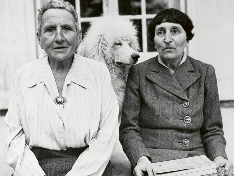 (Original Caption) Gertrude Stein, one of the world's foremost novelists, poses with Alice B Toklas, and her dog, Basket, in front of her home in France. The grand lady of fiction lived in a chateau near Paris during the four years of Nazi occupation, but her identity was kept secret. She has written a new book, part of which Miss Toklas is holding, which will be released for publication shortly.